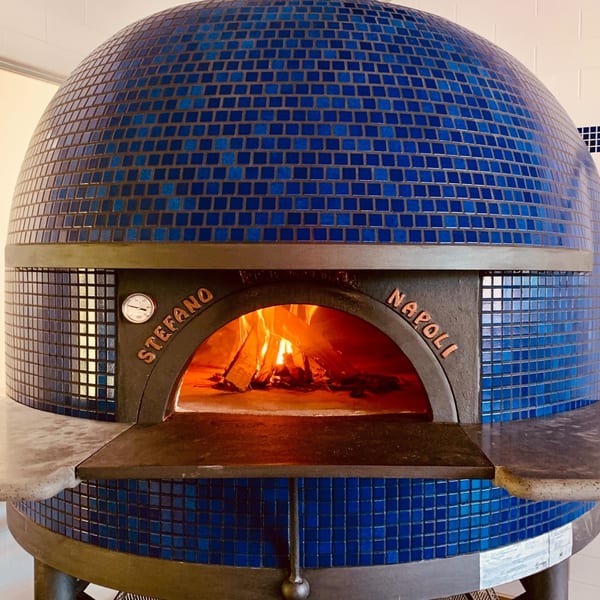 A large, blue tiled Pizzaluté wood-fired oven with a roaring fire inside.